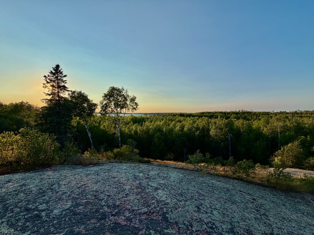 River flowing through a boreal forest landscape at sunset, with birch and conifer trees lining the banks and a clear sky above