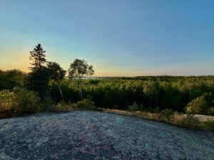 River flowing through a boreal forest landscape at sunset, with birch and conifer trees lining the banks and a clear sky above