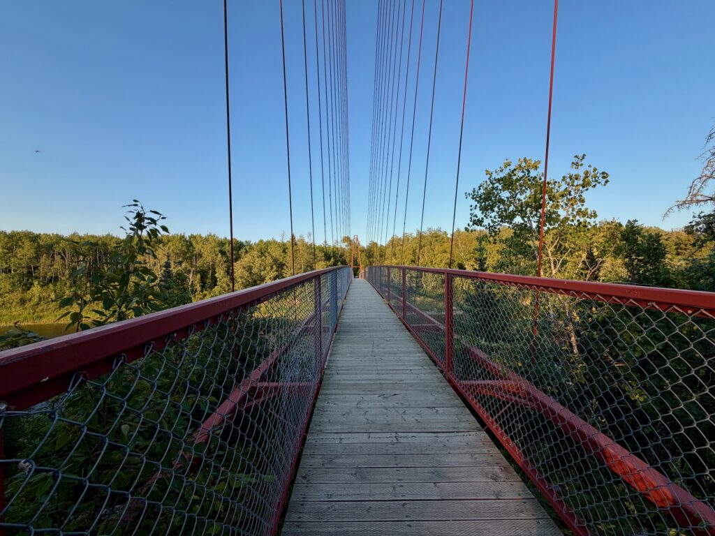 Suspension bridge with red railings and cable supports stretching over a forested landscape under clear blue sky