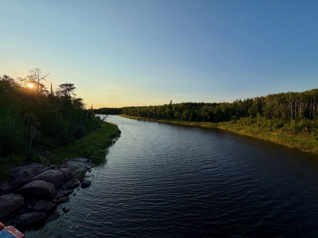 River winding through boreal forest at sunset, with rocky shoreline in foreground and coniferous trees lining both banks under a clear sky