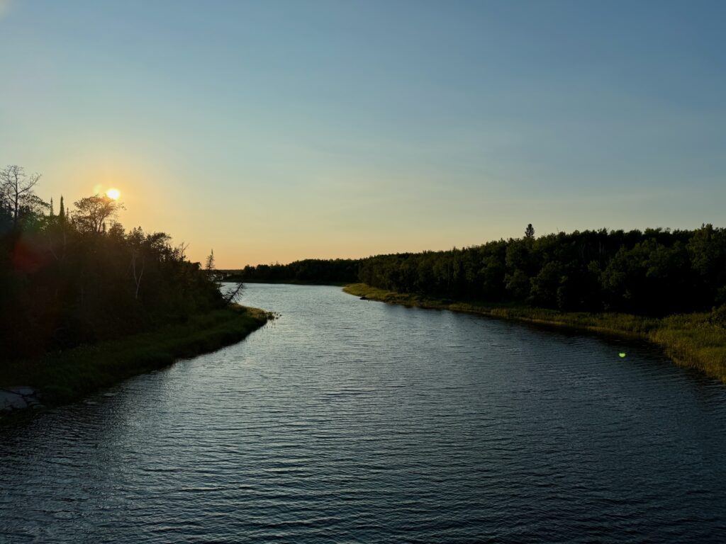 River flowing through a forested landscape at sunset, with coniferous trees lining both banks and the sun low on the horizon casting golden light across the water