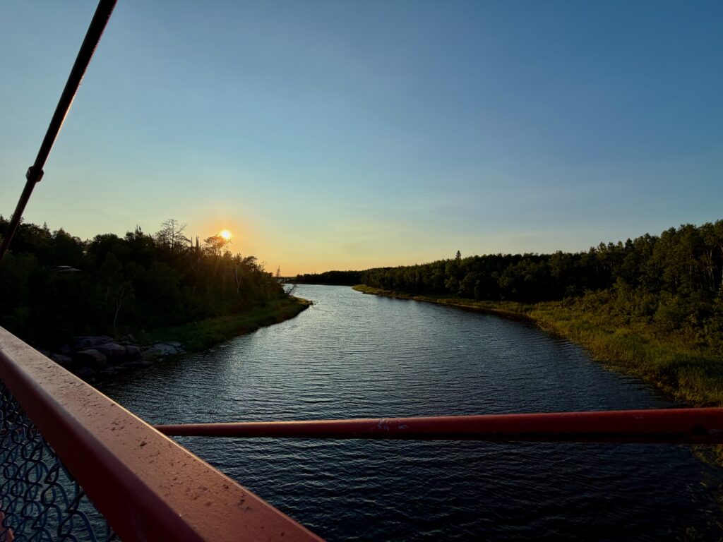 River at sunset viewed from a bridge railing, with forested shorelines on both sides and the sun low on the horizon