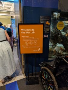 Welcome to the Wet Lab safety sign with orange background displaying rules: rinse hands at sinks inside, listen to an educator before touching, touch only with your pinky finger, and note that the area may not be suitable for children under 4. Vancouver Aquarium logo at bottom