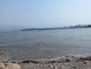 Vancouver skyline viewed across the water from a rocky beach, with sailboats and forested shores visible in the middle distance
