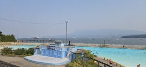 Kitsilano Pool in Vancouver with turquoise water, changing pavilion, sailboats, and container ships on Burrard Inlet under clear sky