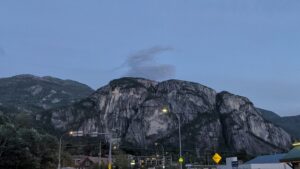 Steep columnar rock formations rising above a small town at dusk, with street lights illuminated and mountains in the background