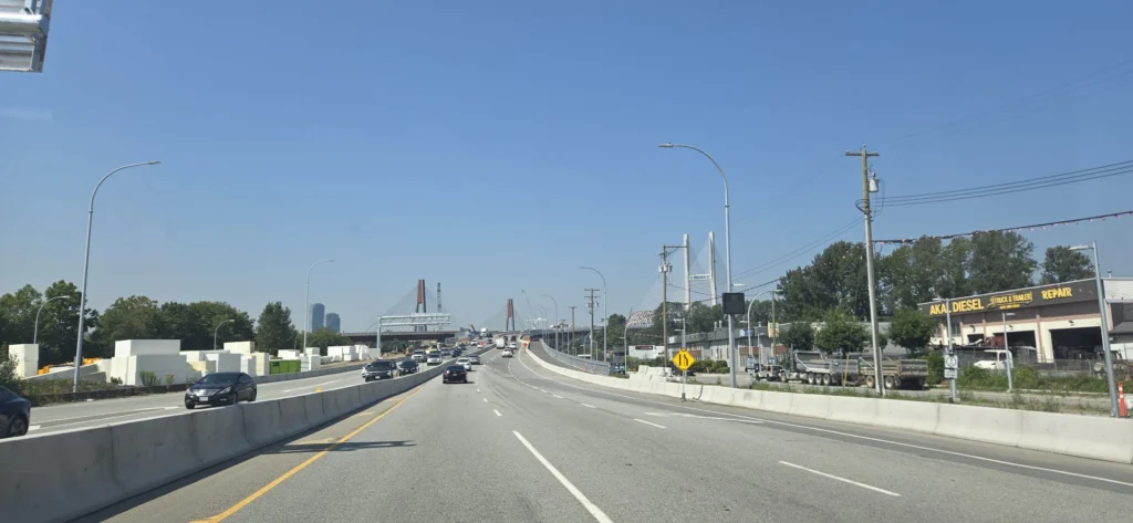 View from a vehicle on a multi-lane bridge with concrete barriers, approaching an iconic white cable-stayed bridge with red towers in the distance under clear blue sky
