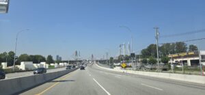 View from a vehicle on a multi-lane bridge with concrete barriers, approaching an iconic white cable-stayed bridge with red towers in the distance under clear blue sky