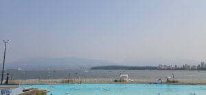 Outdoor swimming pool with bright turquoise water overlooking a bay dotted with sailboats, mountains across the water, and a city skyline under hazy sky
