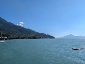 Turquoise lake surrounded by forested mountains under clear blue sky, with a moored sailboat and distant peaks across the water