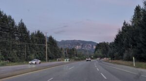 Highway stretching through a forested valley with mountains visible in the distance under a twilight sky, with vehicles traveling in both directions