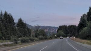 Highway stretching through forested valley with mountains and cliffs visible in the distance under twilight sky