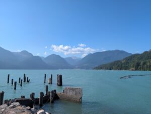 Turquoise lake surrounded by forested mountains and snow-capped peaks, with weathered wooden pilings and a concrete structure in the shallow water