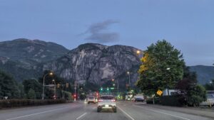 Dusk view from a car driving towards a valley road with traffic lights, street lamps, and forested mountains with columnar basalt cliffs in the background