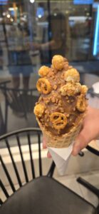 Ice cream cone topped with peanut butter, cereal pieces, and pretzel crisps, held in a hand at a restaurant
