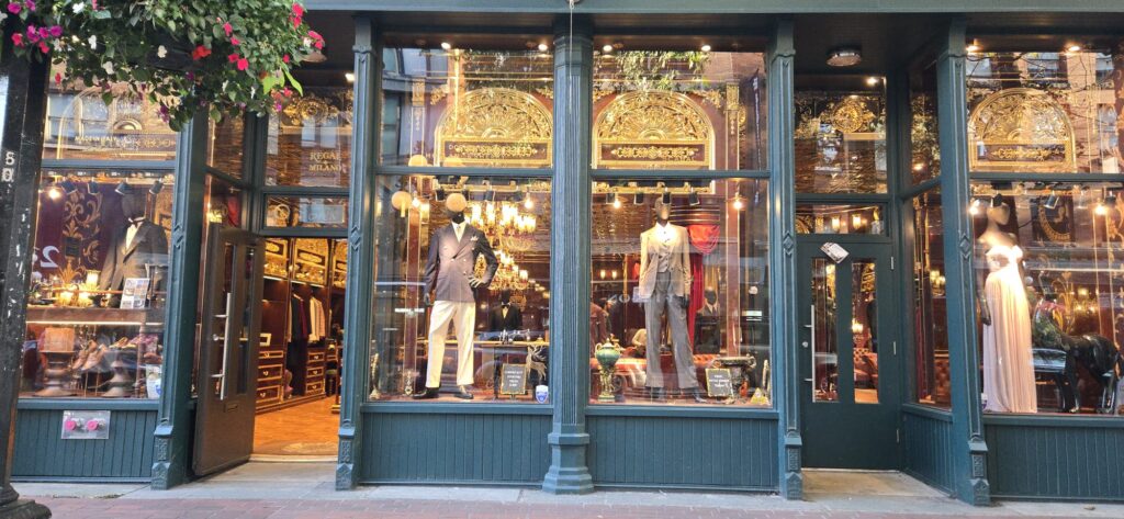 Luxurious fashion boutique storefront with teal-framed windows displaying mannequins in formal attire, ornate gold-trimmed mirrors, warm lighting, and pink flowers above the entrance