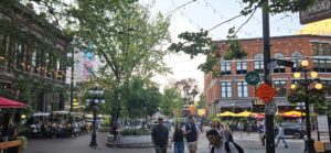 Busy urban plaza with historic brick buildings, overhead string lights, green trees, outdoor seating with coloured umbrellas, and pedestrians walking and dining in a vibrant downtown district