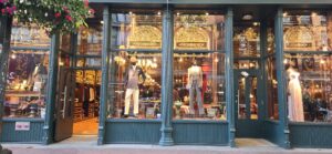 Luxury fashion boutique storefront with large display windows featuring mannequins in formal wear, ornate gold decorative panels, warm lighting, and pink flowers above the entrance