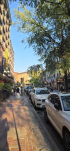 A busy urban street lined with parked cars and pedestrians walking beneath tall trees with autumn foliage. Historic brick buildings and shops line both sides of the street under a clear blue sky