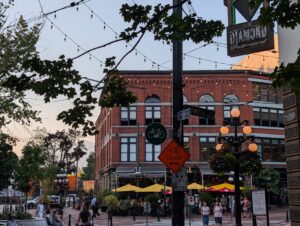 Busy urban plaza with historic red-brick buildings, strung pendant lights, a bicycle detour sign, orange globe street lamps, and pedestrians browsing outdoor market stalls with yellow and red umbrellas