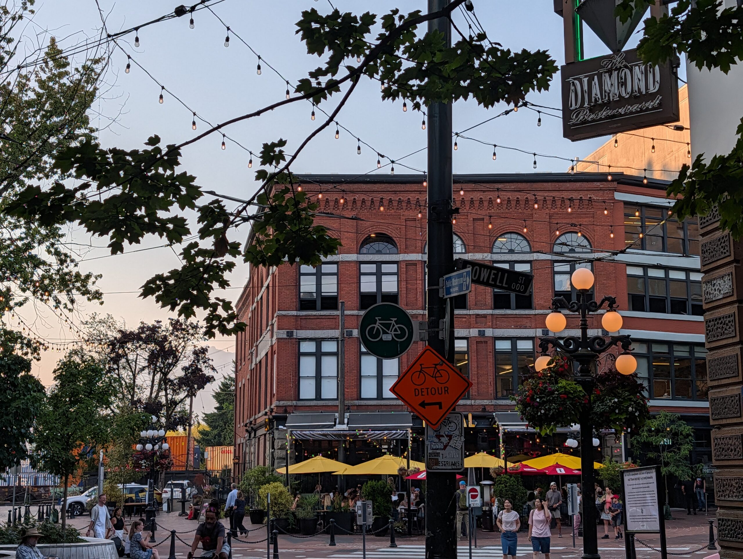 Busy urban plaza with historic red-brick buildings, strung pendant lights, a bicycle detour sign, orange globe street lamps, and pedestrians browsing outdoor market stalls with yellow and red umbrellas