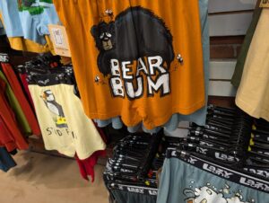 Orange t-shirt featuring a black bear with white text reading "BEAR BUM" and small bee illustrations, displayed on a retail rack with other merchandise