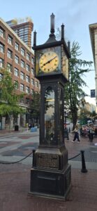 The Gastown Steam Clock, a black cast-iron street clock with a cream-coloured face displaying Roman numerals, stands in a brick plaza surrounded by historic buildings and pedestrians in Vancouver