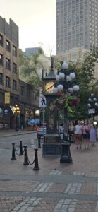 Historic street clock with illuminated globe lamps and hanging flower baskets in an urban pedestrian area, surrounded by brick buildings and modern high-rises, with steam rising in the background