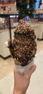 Chocolate-dipped ice cream cone topped with toffee bits and chocolate drizzle, held in a hand over a blurred indoor venue