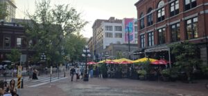 Busy urban plaza with brick and heritage buildings, pedestrians walking, and outdoor seating area with yellow and red umbrellas beneath a covered structure