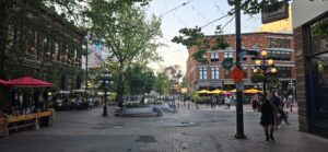 Busy pedestrian plaza with brick heritage buildings, string lights, yellow umbrellas, tree canopy, and people walking and dining at outdoor patios