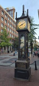 The Gastown Steam Clock, a historic cast-iron street clock with a cream-coloured face and Roman numerals, situated in a public plaza surrounded by brick buildings and pedestrians