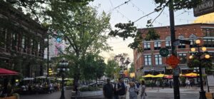 Busy urban plaza with historic brick buildings, tree-lined streets, string lights, and people walking among yellow and red umbrellas