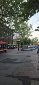 Pedestrian plaza with mature trees, brick buildings, outdoor seating under a red canopy, and people gathered in an urban public square on a summer day
