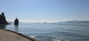Calm coastal waters with rocky outcrops, distant mountains, and cargo ships anchored offshore under clear skies