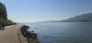 Waterfront promenade with paved pathway along rocky shoreline, people walking, calm bay water, forested hillside on left, and snow-capped mountains with distant city skyline across the water under clear blue sky