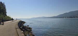 Waterfront seawall pathway with people walking, overlooking calm blue water, distant mountains, and a city skyline across the bay on a clear day