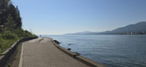 Waterfront pathway along a calm bay with forested shoreline on the left, mountains across the water, and a city skyline visible in the distance