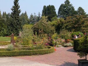 A formal garden with brick pathways, manicured hedges, flowering perennials in pink and red, tall evergreen and conifer trees in the background, and visitors strolling through the landscaped grounds on a clear day