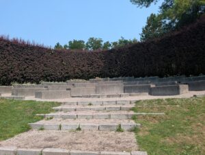 Outdoor amphitheatre with tiered concrete seating, surrounded by dark hedges and trees under clear sky