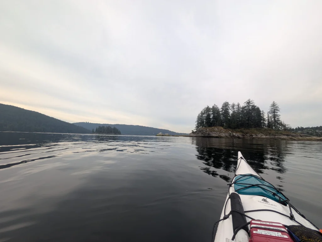 Kayak with teal spray skirt on calm water, forested rocky island in distance, coniferous trees lining distant shoreline under overcast sky
