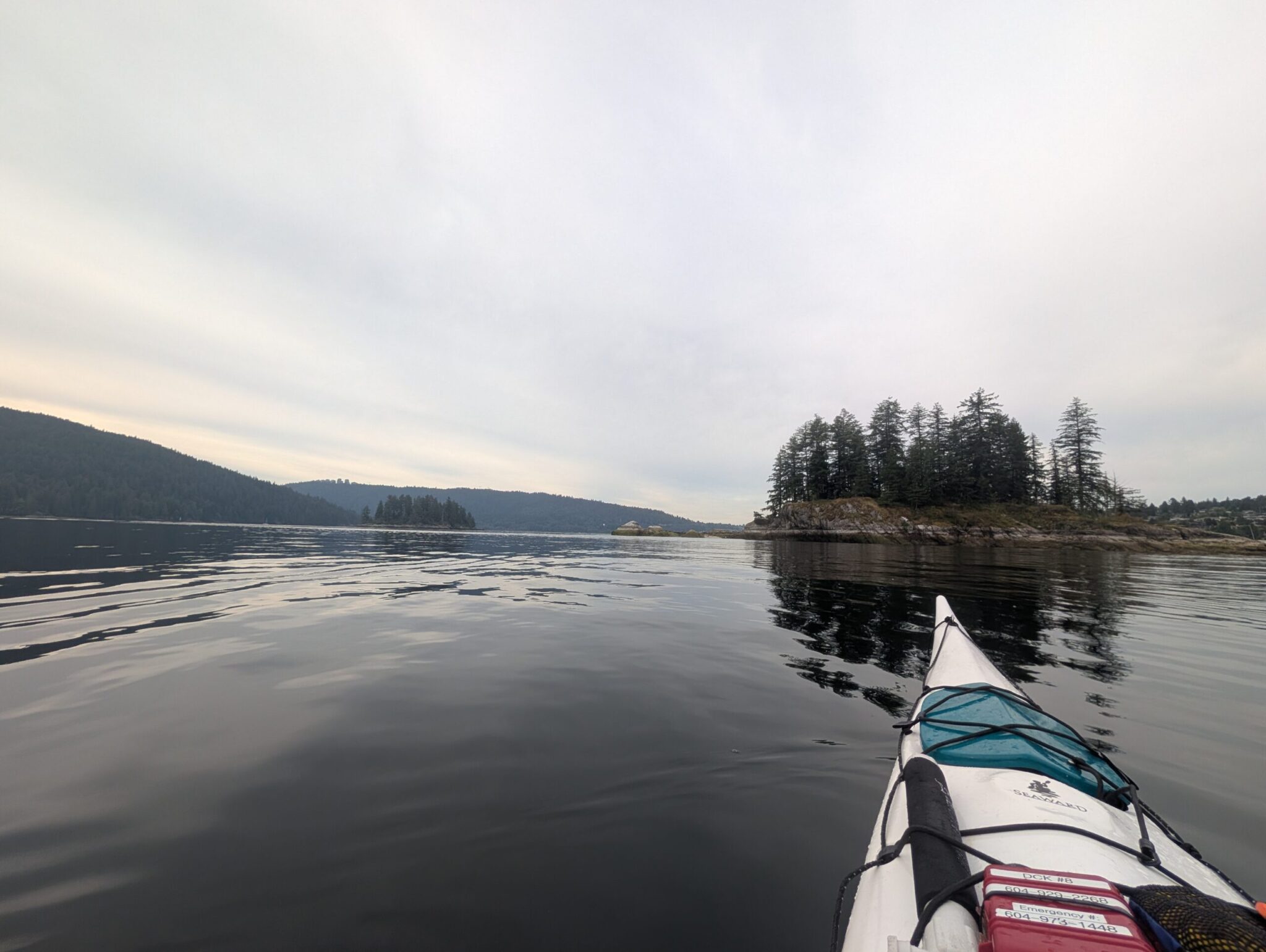 Kayak with teal spray skirt on calm water, forested rocky island in distance, coniferous trees lining distant shoreline under overcast sky
