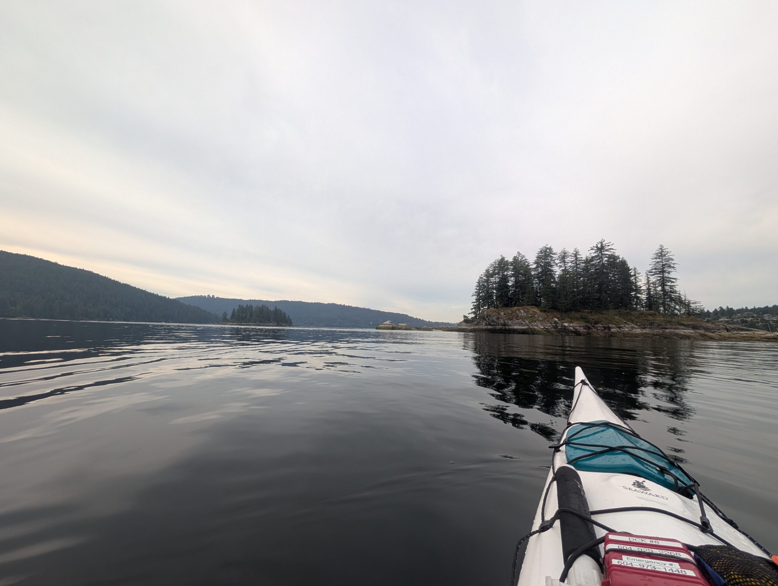 Kayak with teal spray skirt on calm water, forested rocky island in distance, coniferous trees lining distant shoreline under overcast sky