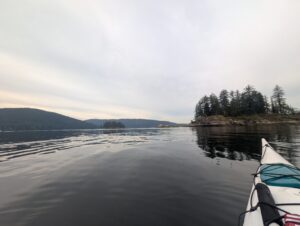 Kayak bow in calm lake waters with forested shoreline and mountains in the distance under overcast sky