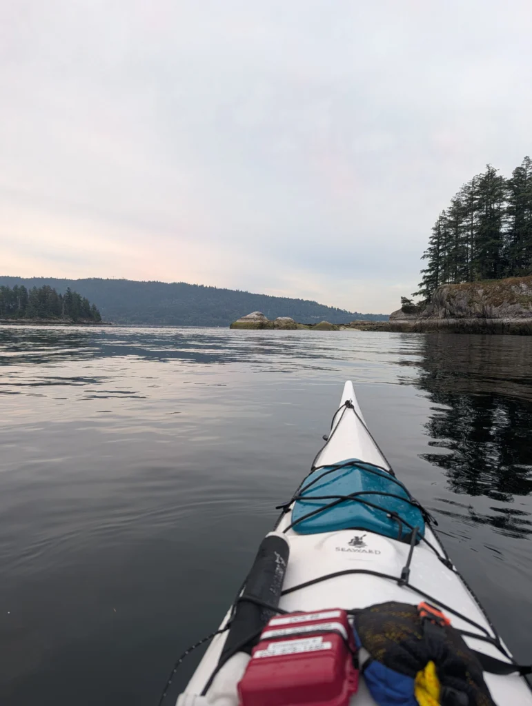 View from the bow of a kayak on calm water, with forested shorelines, rocky islands, and mountains in the distance under an overcast sky