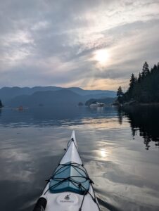 Kayak in calm water on a large lake surrounded by forested mountains under a cloudy sky with distant ferry boat and shoreline cabins