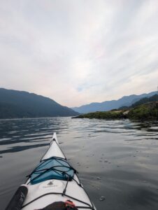 View from a sea kayak on a calm lake surrounded by forested mountains under a cloudy sky