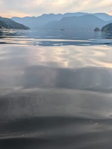 Calm mountain lake at dawn with forested shores and distant peaks shrouded in mist, taken from the water's edge