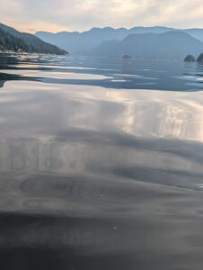 Serene mountain lake at dawn with forested shoreline, distant peaks, and calm water reflecting soft clouds