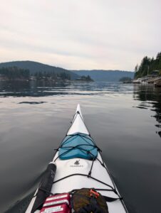 View from the bow of a sea kayak on a calm lake surrounded by forested mountains and distant shoreline cottages